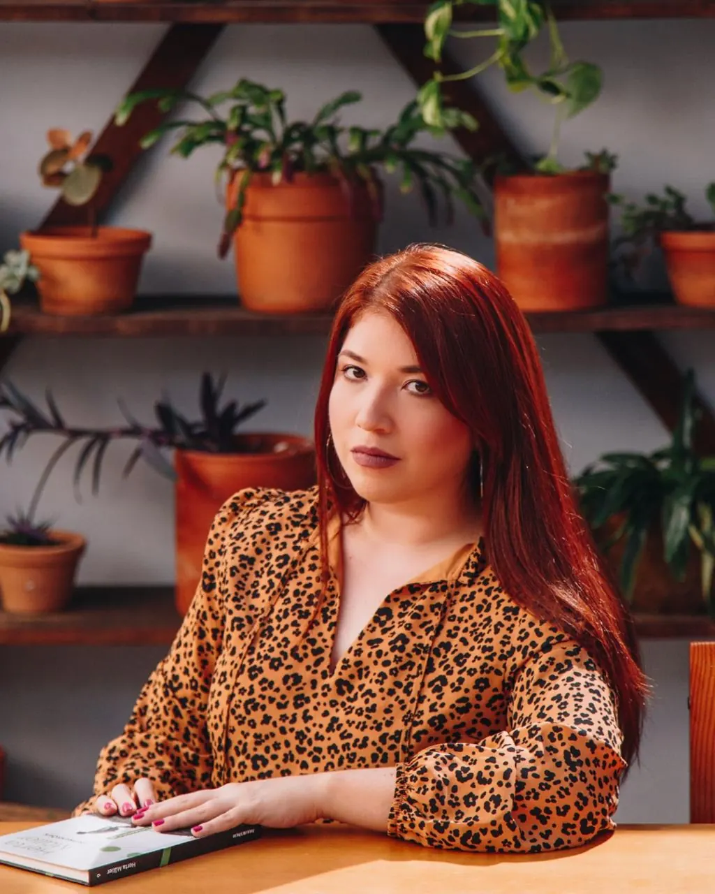 Michelle Santiago sentada à mesa, com um livro, diante de uma estante com plantas